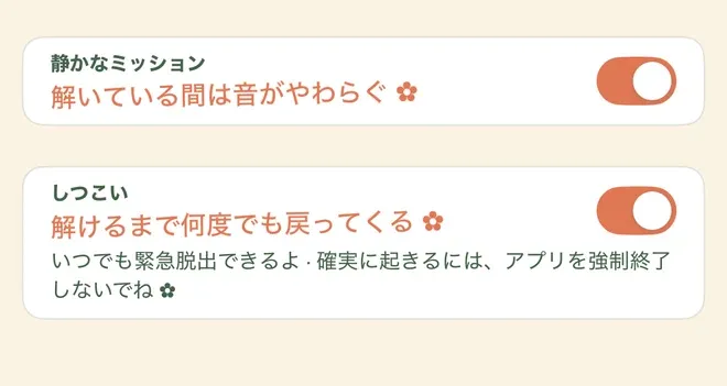 アラームの設定が縦に二つ並んだ画面。上は「静かなミッション」で、値は「解いている間は音がやわらぐ」、右側のスイッチはオン。下は「しつこい」で、値は「解けるまで何度でも戻ってくる」、こちらもスイッチはオン。その下に「いつでも緊急脱出できるよ」と「確実に起きるには、アプリを強制終了しないでね」が中黒でつながった案内文が一行入る。値の行と案内文の末尾には小さな花の飾りが付く。