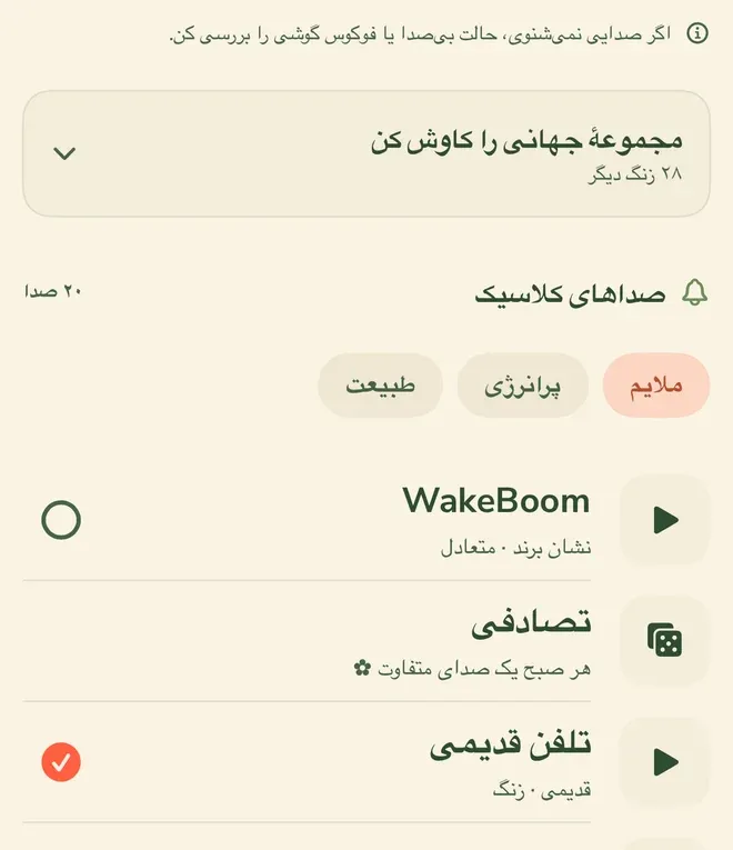 فهرست صداهای زنگ. بالای صفحه یادداشتی با نشان اطلاعات: «اگر صدایی نمی‌شنوی، حالت بی‌صدا یا فوکوس گوشی را بررسی کن.» زیر آن کارت «مجموعهٔ جهانی را کاوش کن» با زیرنویس «۲۸ زنگ دیگر»، بعد سرتیتر «صداهای کلاسیک» با شمارش «۲۰ صدا» و سه برچسب گروه: «ملایم» که انتخاب شده، «پرانرژی» و «طبیعت». پایین‌تر سه ردیف صدا: «WakeBoom» با توضیح «نشان برند · متعادل»، «تصادفی» با توضیح «هر صبح یک صدای متفاوت»، و «تلفن قدیمی» با توضیح «قدیمی · زنگ» که تیک نارنجی انتخاب کنارش نشسته. ردیف اول و سوم دکمهٔ پخش دارند و ردیف «تصادفی» به‌جایش نشان تاس دارد.