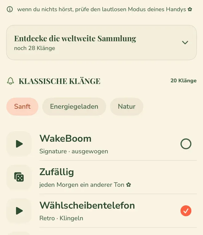 Die Klangauswahl eines Weckers: oben der Hinweis „wenn du nichts hörst, prüfe den lautlosen Modus deines Handys“, darunter die zugeklappte Karte „Entdecke die weltweite Sammlung“ mit „noch 28 Klänge“ und der Abschnitt „KLASSISCHE KLÄNGE“ mit der Angabe „20 Klänge“. Darunter die drei Gruppen „Sanft“ als ausgewählte, „Energiegeladen“ und „Natur“, und die ersten Einträge der Liste: „WakeBoom“ mit „Signature · ausgewogen“, „Zufällig“ mit „jeden Morgen ein anderer Ton“ und das ausgewählte „Wählscheibentelefon“ mit „Retro · Klingeln“.